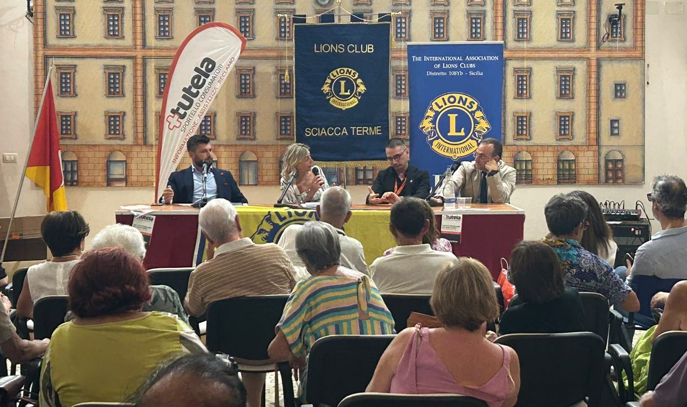 Incontro pubblico in una sala con un tavolo di relatori, il banner dello sportello +Tutela e gli stendardi del Lions Club Sciacca Terme, davanti a un pubblico di cittadini