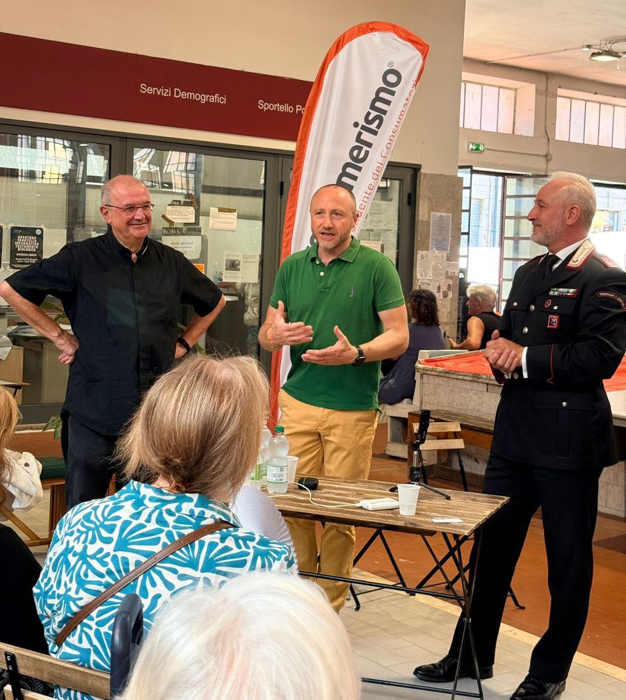 Luigi Gabriele parla a un gruppo di cittadini durante un incontro pubblico, accanto a una bandiera di Consumerismo