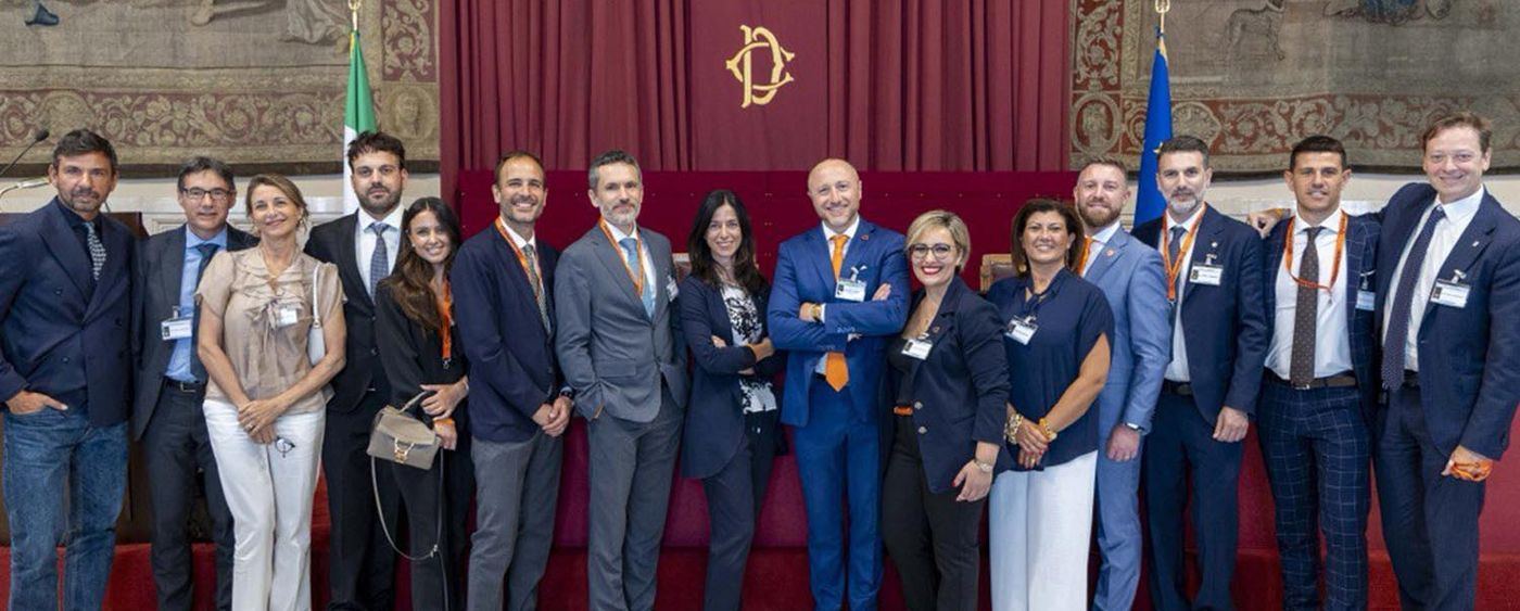 Gruppo di persone in posa in una sala istituzionale, con arazzi alle pareti e le bandiere italiana ed europea